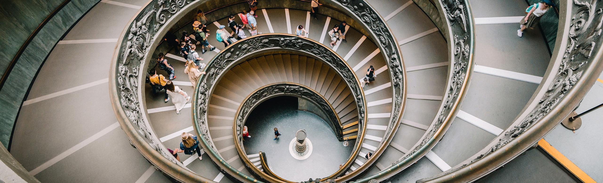 Spiral staircase with visitors – architecture, perspective, flow, heritage Top-down view of a monumental spiral staircase with ornate railing, filled with descending people.