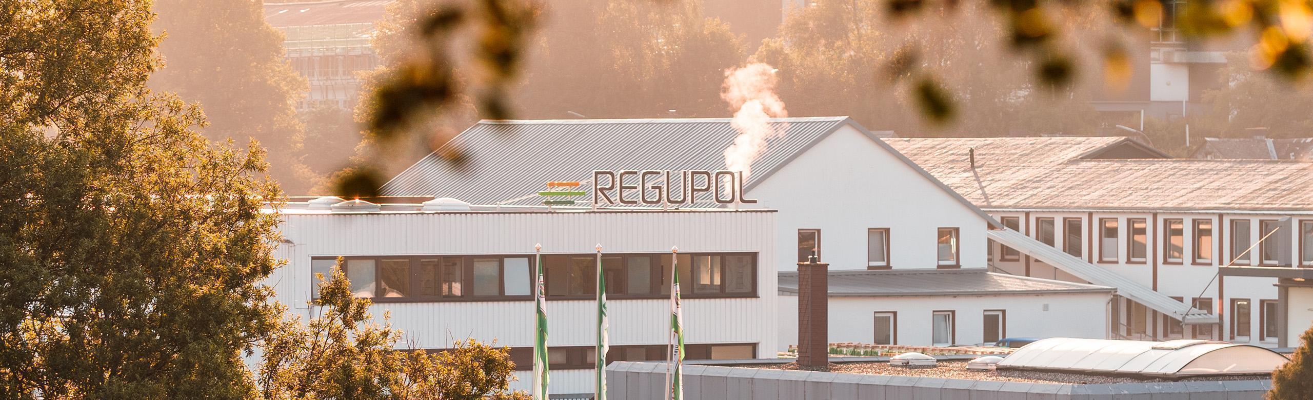 Exterior view of a REGUPOL production site in early morning light. The white industrial building displays the REGUPOL logo on the roof, with steam rising from a chimney. Trees and green REGUPOL flags appear in the foreground.