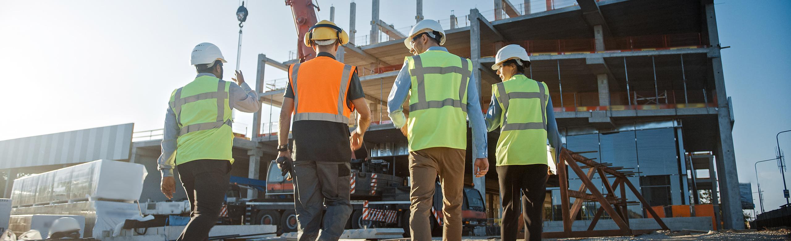Four people wearing safety vests and helmets walk across an active construction site. In the background is a partially constructed concrete building, with crane and equipment visible in bright sunlight.