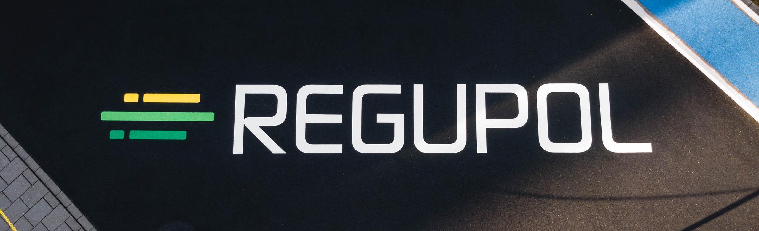REGUPOL logo on rubber granulate – brand, sports flooring, recycling, eco REGUPOL logo in white lettering on black rubber granulate surface, with green and yellow brand bars on the left.